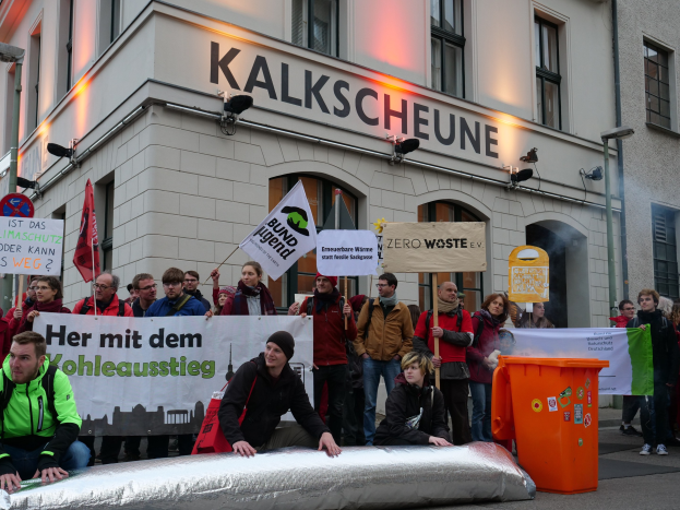 Eine Gruppe von Menschen mit Schildern und Plakaten vor einem Gebäude bei einer Demonstration in Deutschland, mit zwei Menschen im Vordergrund und einem Müllcontainer auf der rechten Seite.