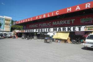 Der Porac Public Market in Manila, Philippinen, zeigt eine belebte Szene mit Fahrzeugen auf der Straße, zahlreichen Ständen, umgeben von Gebäuden und Bäumen unter einem klaren blauen Himmel.