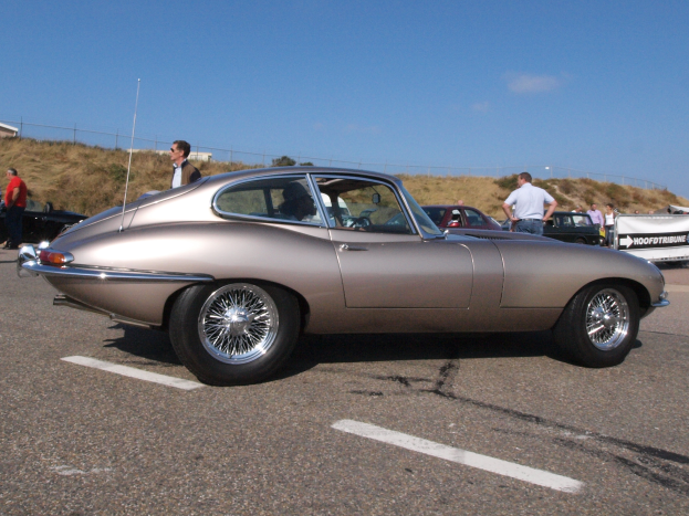Ein silberfarbenes Auto auf einem Parkplatz mit anderen Autos, Menschen in der Nähe, eine Fahne, Masten, ein Zaun, Gras, ein Gebäude und ein bewölkter Himmel.