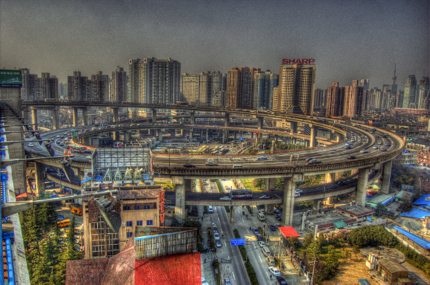 Eine Stadtansicht mit einer zentralen Ringstraße mit Fahrzeugen, einer unteren Straße mit Autos und Fahrrädern, zahlreichen hohen Gebäuden im Hintergrund, einem Himmel oben, Bäumen links unten und kleinen Häusern, die über die Stadt verteilt sind.