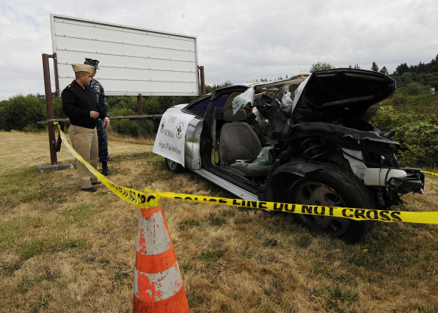 Ein Polizist in Uniform steht neben einem schwer beschädigten Auto am Straßenrand mit eingedellter Haube und zersplitterten Fenstern unter einem bewölkten Himmel.