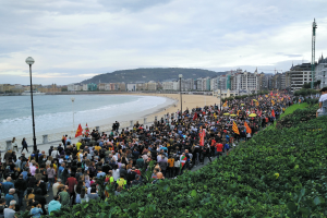 Eine große Gruppe von Menschen steht auf einem Strand in Nizza, Frankreich, hält Fahnen und protestiert gegen Sparmaßnahmen, mit Pflanzen auf der rechten Seite, Laternenpfählen, Gebäuden, Bäumen, Bergen und einem klaren blauen Himmel im Hintergrund.
