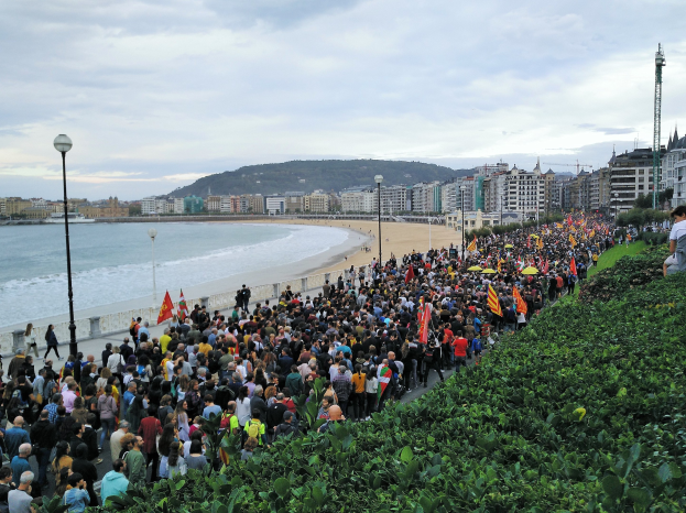 Eine große Gruppe von Menschen steht auf einem Strand in Nizza, Frankreich, hält Fahnen und protestiert gegen Sparmaßnahmen, mit Pflanzen auf der rechten Seite, Laternenpfählen, Gebäuden, Bäumen, Bergen und einem klaren blauen Himmel im Hintergrund.