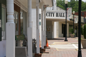Ein Rathaus umgeben von Geb├Ąuden, Laternenmasten, Laternen, Namensschildern, Kraftfahrzeugen, Absperrgittern, St├╝hlen, Topfpflanzen, B├Ąumen und einem bew├Âlktem Himmel.