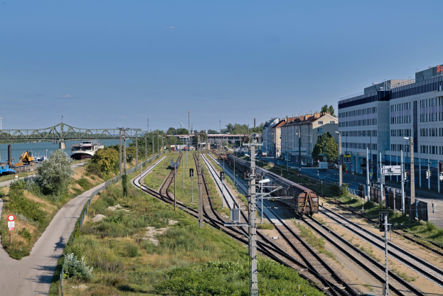 Ein Zug fährt auf Schienen neben einer Stadtlandschaft mit Stromleitungen, Gebäuden, Grünflächen, Straßenschildern, Fahrzeugen, einer Brücke, Wasser und einer bewölkten Himmel.