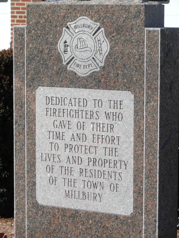 Gedenkstein mit einer Plakette, gewidmet an Feuerwehrleute in Millbury, vor einem Backsteingebäude mit umliegenden Pflanzen und Bäumen.