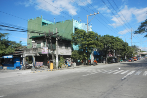 Eine Stadtstraße mit einem grünen Gebäude, fahrende Fahrzeuge, Fußgänger auf dem Gehweg und Strommasten mit Drähten unter einem bewölkten Himmel.