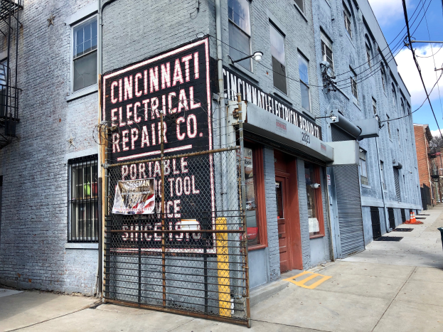 Das Gebäude der Cincinnati Electrical Repair Co. mit einem Schild, einem Metallzaun, einem Müllcontainer, Pfählen, Drähten, Bäumen und einem bewölkten Himmel.