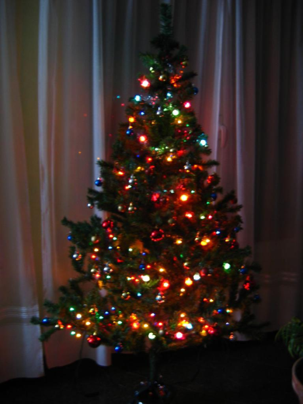 Ein mit Lichtern geschm├╝ckter Weihnachtsbaum steht vor einem Vorhang und einem Topf.