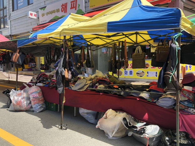 Ein Straßenmarkt mit Zelten, die Taschen, Hüte und andere Artikel präsentieren, entlang einer Straße mit Gebäuden, die Fenster und Texttafeln aufweisen.