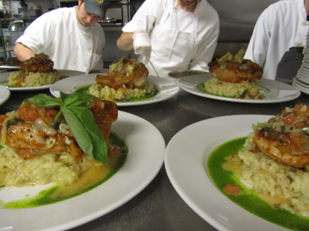 Drei Köche in einer Küche mit Mützen und Handschuhen bereiten Essen auf Tellern zu, die mit verschiedenen Gegenständen gefüllt sind, mit Regalen voller Vorräte im Hintergrund.