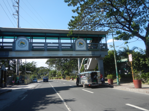Ein Bus fährt eine von Bäumen gesäumte Straße mit einer Brücke im Hintergrund entlang, mit Text und Logos auf Schildern, Strommasten mit Drähten, Laternenmasten und einigen Fußgängern auf dem Gehweg unter einem bewölkten Himmel.