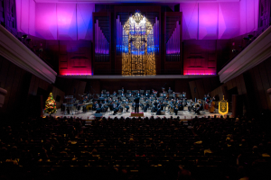 Ein großer Saal voller Menschen, die ein Konzert anschauen, mit Musikern auf der Bühne und anderen im Publikum, einem Weihnachtsbaum im Hintergrund und Wänden mit Lichtern und anderen Gegenständen geschmückt.