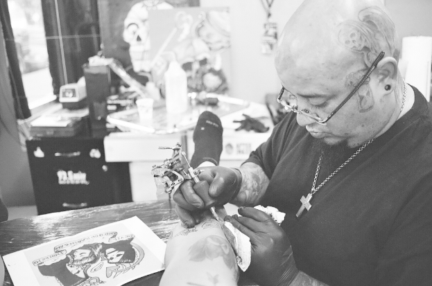 Ein Schwarz-Weiß-Bild eines Mannes in einem schwarzen T-Shirt, Brille und einer Kette um den Hals, der ein Bein mit einer Maschine tätowiert, mit einem Papier auf der Oberfläche und einem unscharfen Hintergrund mit einer Flasche, Tasse und anderen Gegenständen.
