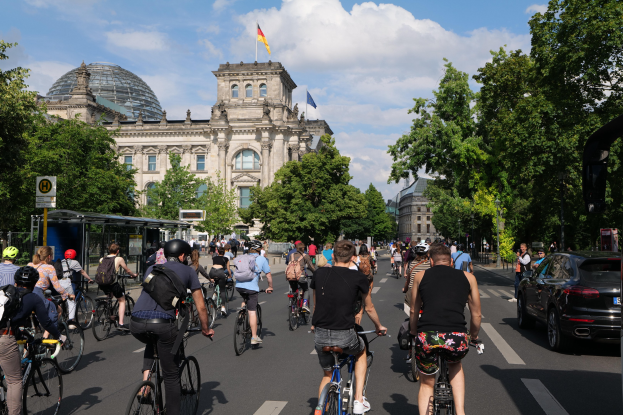 Gruppe von Menschen, die auf Fahrrädern eine von Bäumen gesäumte Straße in Berlin, Deutschland, entlangfahren, mit einer Bushaltestelle auf der rechten Seite und einer Flagge, die auf einem Gebäude weht, unter einem bewölkten Himmel.