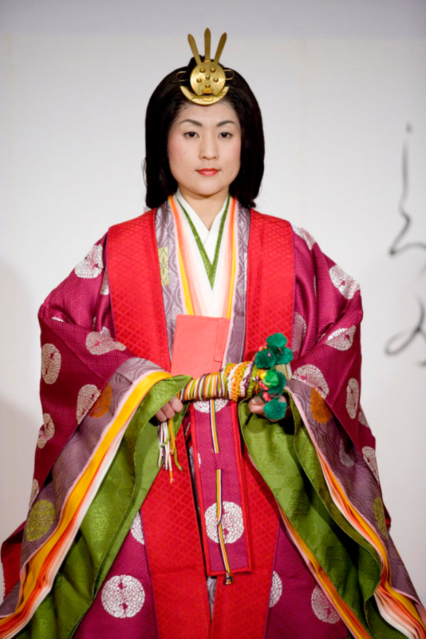 Eine Frau in einem traditionellen japanischen Kimono, mit einem leuchtend roten und grünen Kleid mit aufwändigen Mustern, steht selbstbewusst auf einem Laufsteg, ihr Haar ist ordentlich zu einem Dutt gebunden.