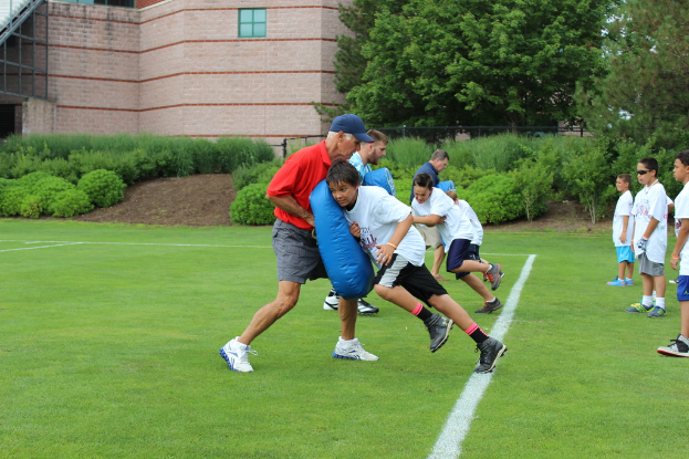 Mehrere Kinder versuchen, durch eine Gruppe von Männern zu gelangen, die ihnen den Weg versperren, mit weiteren Kindern davor und Bäumen, einem Gebäude und Büschen im Hintergrund.
