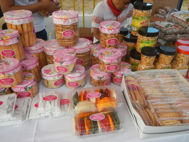 Eine Gruppe von Menschen um einen Tisch versammelt, auf dem verschiedene verpackte Lebensmittel ausgebreitet sind, mit einem Geländer und einer Straße im Hintergrund.