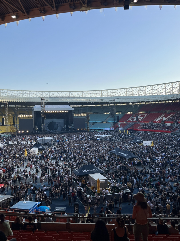 Große Menschenmenge auf Sitzplätzen und Stehplätzen in einer Halle mit Deckenbeleuchtung und sichtbarem Himmelhintergrund, was auf ein Konzert in der O2 Arena in London hindeutet.