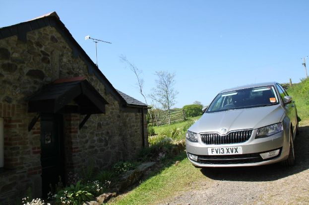 Eine Szene mit einem Haus und einem Zaun auf der linken Seite und einem Auto, Pflanzen, einem Strommast und Gras auf der rechten Seite unter einem sonnigen Himmel.