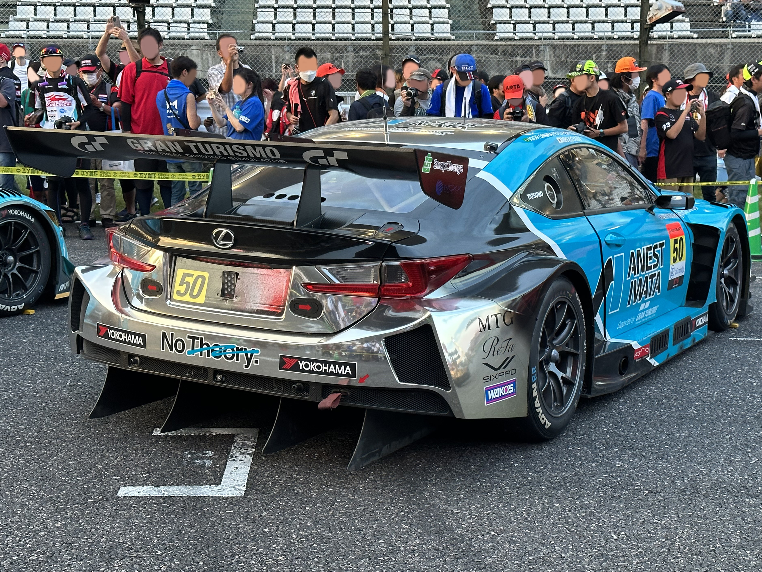 Ein Lexus RC F GT3 Rennwagen auf der Straße, umgeben von Menschen in Möppen und Schutzbrillen, einige halten Kameras und Telefone, mit Maschendrahtzaun und Stühlen im Hintergrund.