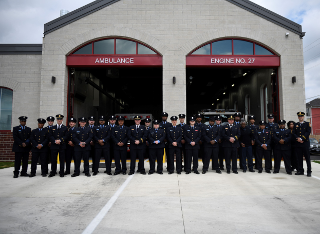 Feuerwehrleute in Uniform und Mützen stehen vor einer Feuerwache mit Fahrzeugen, Gebäuden und einem klaren Himmel im Hintergrund.