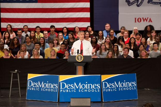 Ein Mann in einem weißen Hemd und roter Krawatte steht hinter einem Pult mit drei Texttafeln, einem Hocker mit Mikrofon und einer sitzenden Zuhörerschaft im Hintergrund, mit einer Flagge und zusätzlichem Text.