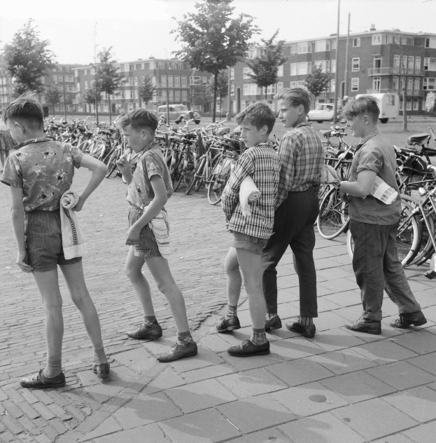 Schwarz-weißes Foto einer Gruppe junger Jungs, die nebeneinander auf einem Bürgersteig stehen, umgeben von Fahrrädern, Fahrzeugen, Bäumen, Pfählen und Gebäuden im Hintergrund.