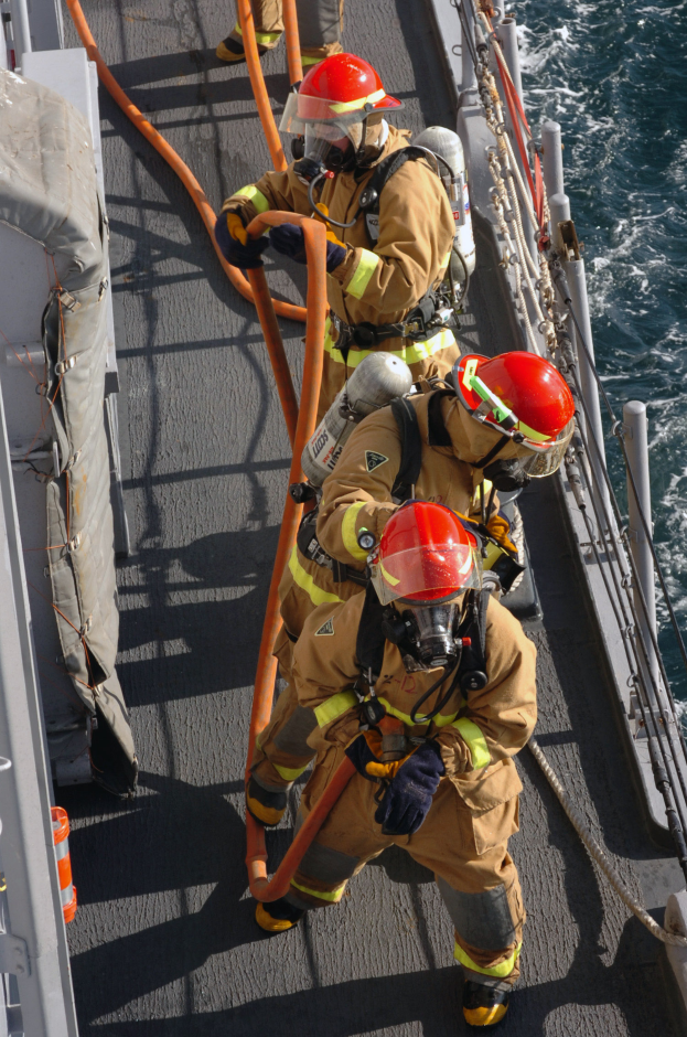 Feuerwehrleute mit Helmen und Handschuhen, die Rohre tragen, gehen auf dem Deck eines Schiffes mit Seilen und Stangen auf der rechten Seite und Wasser im Hintergrund.