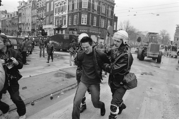 Ein Schwarz-Weiß-Bild eines Mannes, der von einem Polizeibeamten auf der Straße eskortiert wird, mit einer Gruppe von Menschen mit Kameras, Fahrzeugen, Schildern, Straßenlaternen, Verkehrsampeln, Kabeln, Bäumen, Gebäuden mit Fenstern und einer bewölkten Himmel.
