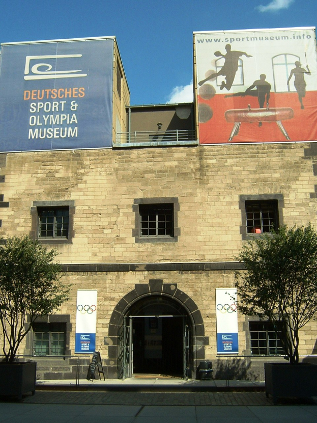 Das Deutsche Sport & Olympische Museum in Berlin, Deutschland, ein Gebäude mit Fenstern und einer Tür, umgeben von Bäumen und Bannern mit Text und Bildern, unter einem bewölkten Himmel.