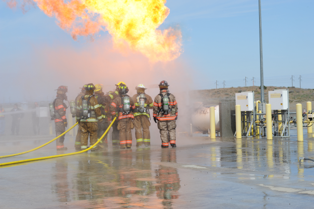Eine Gruppe von Feuerwehrleuten in Helmen, die Rohre halten, steht vor einem großen Feuer mit verstreuten Pfählen, Drähten und anderen Gegenständen auf dem Boden unter einem bewölkten Himmel.
