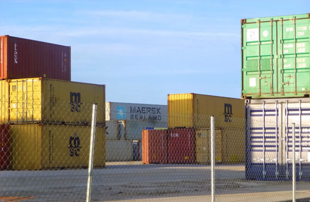 Stapel von Containern hinter Maschendrahtzaun mit Straße und wolkenverhangenem Himmel im Hintergrund.