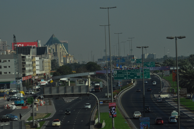 Eine Luftaufnahme einer Stadt mit Gebäuden, Türmen, Kraftfahrzeugen, Werbetafeln, Schildern, Straßen, Bäumen, Straßenlaternen, Menschen, Absperrgittern und Informationsschildern unter einem klaren Himmel.