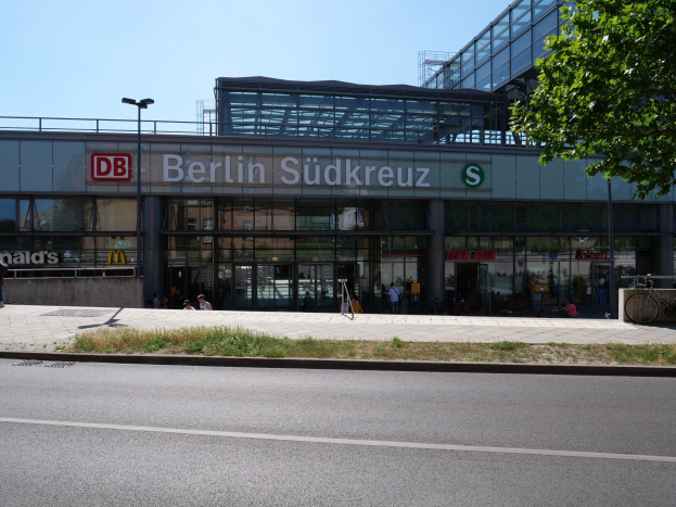 Gro├čer gl├Ąserner Berliner Südkreuz-Bahnhof mit sichtbarem Text, umgeben von Stra├čenm├Âbeln, Fahrzeugen, Passanten, Fahrr├Ądern, B├Ąumen und einem klaren Himmel.