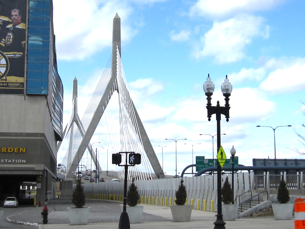 Die Zakim Bridge in Boston, Massachusetts, mit einem Baugerüst auf der linken Seite, Straßenlaternen, Ampeln, Schildern, Topfpflanzen, einem Hydranten und Fahrzeugen auf der rechten Seite, unter einem bewölkten Himmel.