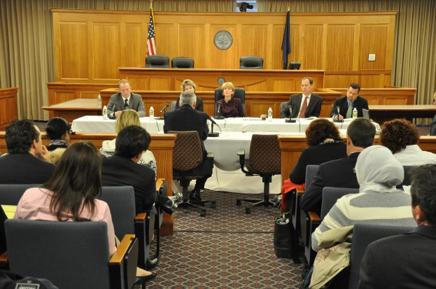 Ein Versammlungsraum mit einer Gruppe von Menschen, die sitzen, zwei ausländische Flaggen, ein orangefarbener Schreibtisch mit Wasserflaschen und einem Mikrofon.