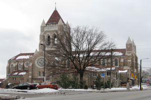 Eine gro├če Kirche mit einem Turm ist von Geb├Ąuden, Stra├čenlaternen, Strommasten, Stromkabeln, Kraftfahrzeugen auf einer schneebedeckten Stra├če, B├Ąumen und einem bew├Âlktem Himmel umgeben.