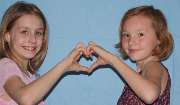 Zwei Kinder lächeln und bilden ein Herz mit ihren Händen vor einer Wand.