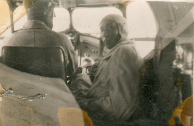 Altes Schwarz-Weiß-Foto von zwei Männern im Cockpit eines Flugzeugs, einer sitzt im Pilotensitz und trägt eine Uniform, der andere steht neben ihm, beide haben ernste Gesichter.