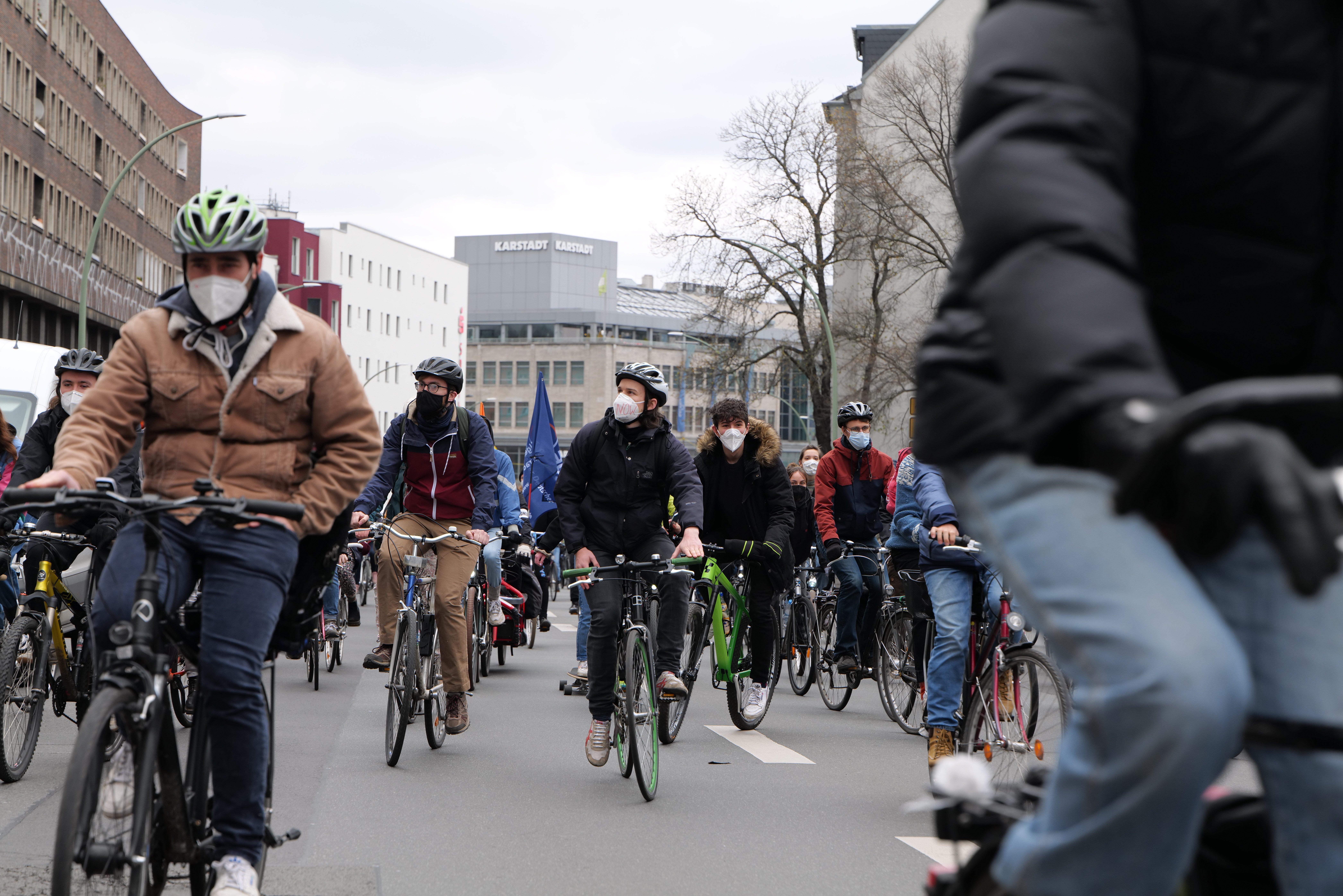 Eine Gruppe von Menschen mit Helmen und Handschuhen fährt auf Fahrrädern eine von Bäumen gesäumte Straße in Berlin, Deutschland, entlang, wobei Gebäude und ein geparktes Fahrzeug im Hintergrund zu sehen sind.