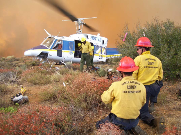 Eine Gruppe von Feuerwehrmännern in gelben Uniformen und roten Helmen steht neben einem Hubschrauber auf einem grasigen Feld, mit Pflanzen, Bäumen und einem raucherfüllten Himmel im Hintergrund.