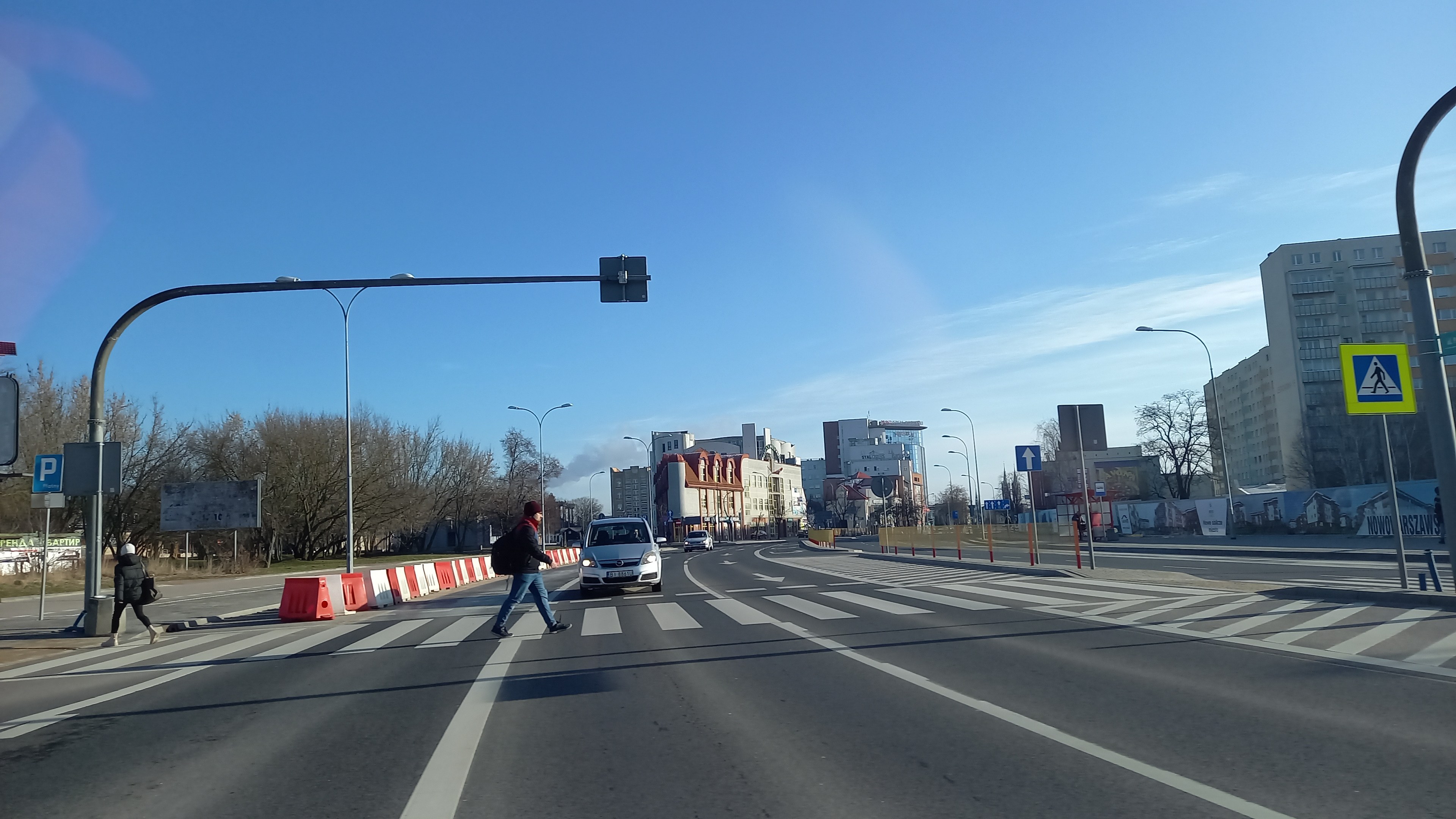 Ein Mann überquert die Straße an einer Ampel mit Fahrzeugen, Fußgängern, Verkehrskegeln, Schildern, Straßeninfrastruktur, Bäumen, Gebäuden und einem bewölkten Himmel im Hintergrund.