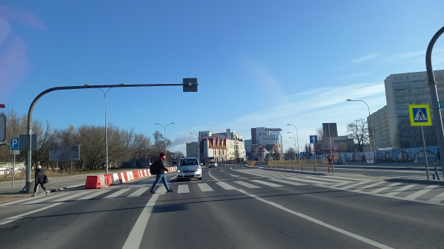 Ein Mann überquert die Straße an einer Ampel mit Fahrzeugen, Fußgängern, Verkehrskegeln, Schildern, Straßeninfrastruktur, Bäumen, Gebäuden und einem bewölkten Himmel im Hintergrund.