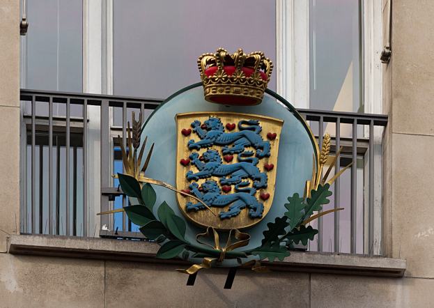 Das Wappen der britischen Königsfamilie wird auf der Außenseite eines Gebäudes zusammen mit einem Kronen- und Blätter-Logo gezeigt, mit Fenstern und Metallgittern im Hintergrund.