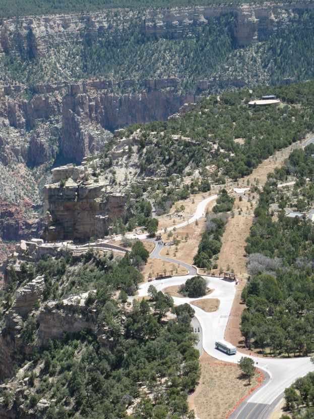 Ein Fahrzeug fährt auf einer Straße umgeben von Bäumen, mit Hügeln im Hintergrund und einem Gebäude auf der rechten Seite.