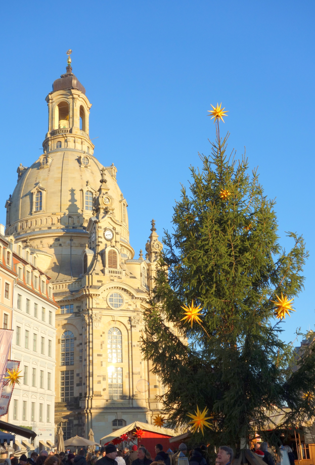 Ein belebter Weihnachtsmarkt in Dresden, Deutschland, mit einem großen Weihnachtsbaum vor einer Kirche, vielen Menschen drumherum und einer Fahne mit Text auf der linken Seite, unter einem sichtbaren Himmel.