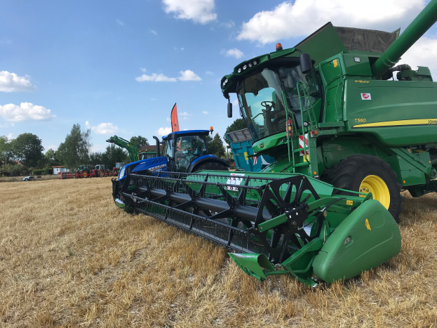 Ein John-Deere-Mähdrescher auf einem trockenen Grasfeld mit Bäumen und einem bewölkten Himmel im Hintergrund.