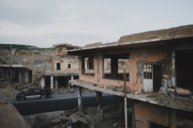 Ein Lastwagen fährt auf einer Straße neben einem zerstörten Gebäude mit zerbrochenen Wänden und verstreuter Trümmer, mit anderen Gebäuden, Polen, Bäumen, einem Hügel und dem Himmel im Hintergrund.