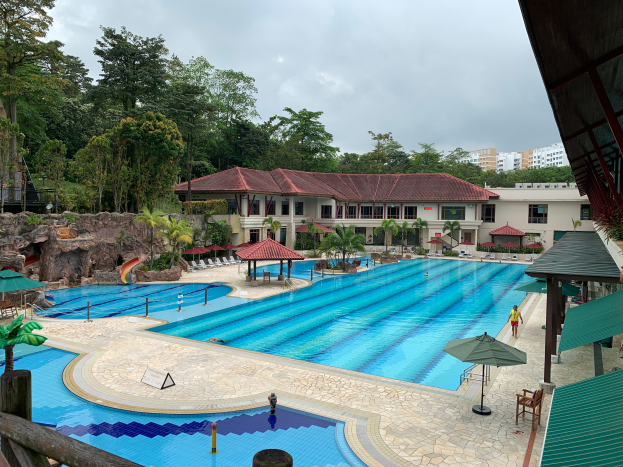 Ein großes Schwimmbad im Ritz-Carlton, Kota Kinabalu, voller Menschen, umgeben von Schirmen, Stühlen, Tischen, Pflanzen, Bäumen, Felsen und Gebäuden, mit einem Schuppen auf der rechten Seite und dem Himmel im Hintergrund.